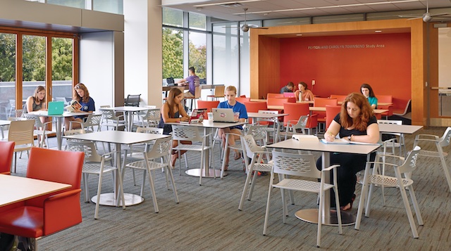 students sit and work at a number of tables in a common study area